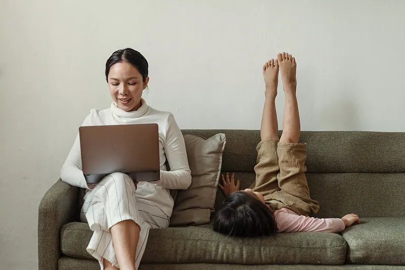 Una mujer trabaja en su computadora en el sillón mientras una niña juega a su lado.