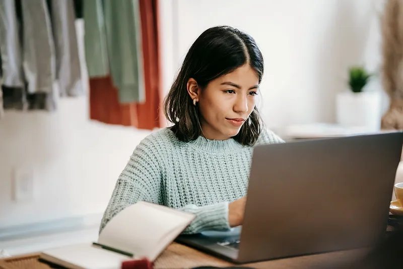 Una mujer joven estudia en una computadora portátil con un cuaderno abierto.